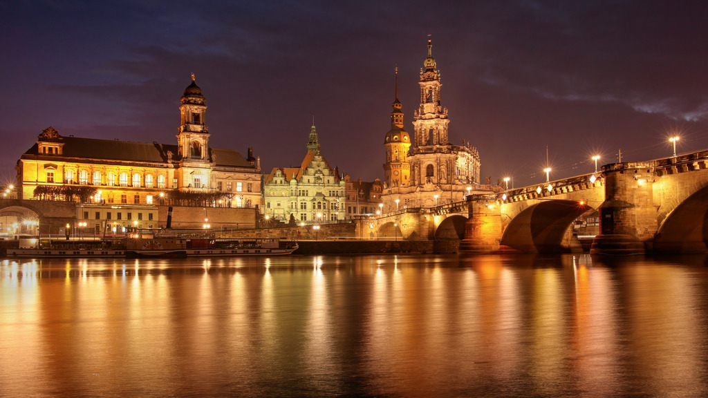 Dresden im Abendlicht - Hofkirche und Augustusbrücke | Dieses Panorama-Wandbild zeigt Dresden von einer seiner prachtvollsten Seiten. Im warmen Licht der Nacht erstrahlt die Hofkirche in voller Schönheit, flankiert von der historischen Augustusbrücke. Die Spiegelungen auf der ruhigen Elbe verleihen der Szene eine malerische Tiefe. Architektur, Wasser und Licht vereinen sich zu einem harmonischen Stadtbild, das Ruhe und Eleganz ausstrahlt. Ideal für alle, die Dresden lieben oder ihren Räumen eine kultivierte und stimmungsvolle Note verleihen möchten.