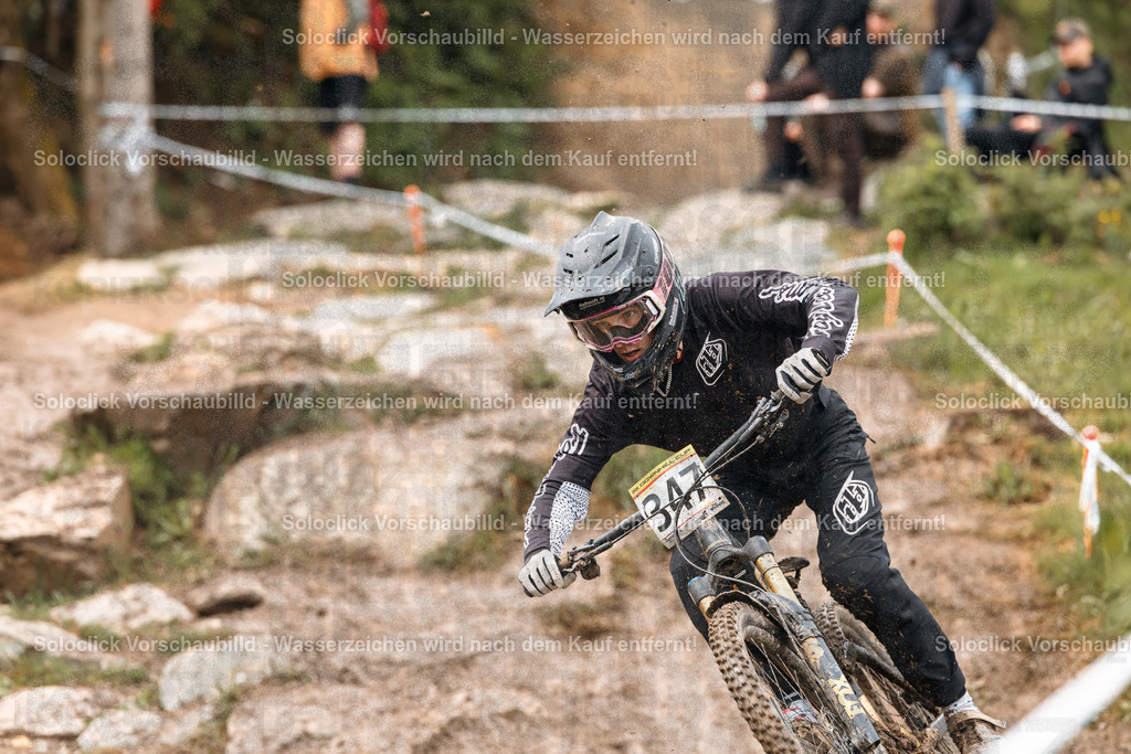 Mountainbike-Action im Wald | Ein Mountainbiker rast auf einem anspruchsvollen Trail durch einen dichten Wald. Er trägt einen schwarzen Helm mit Visier, eine dunkle Jacke und Handschuhe. Er fährt ein Full-Suspension-Mountainbike mit breiten Reifen und einer roten Federgabel. Der Trail ist mit Steinen und Wurzeln übersät und wird von weißen Markierungspfosten gesäumt. Im Hintergrund sind weitere Zuschauer und Bäume zu sehen. - Realisiert mit Pictrs.com