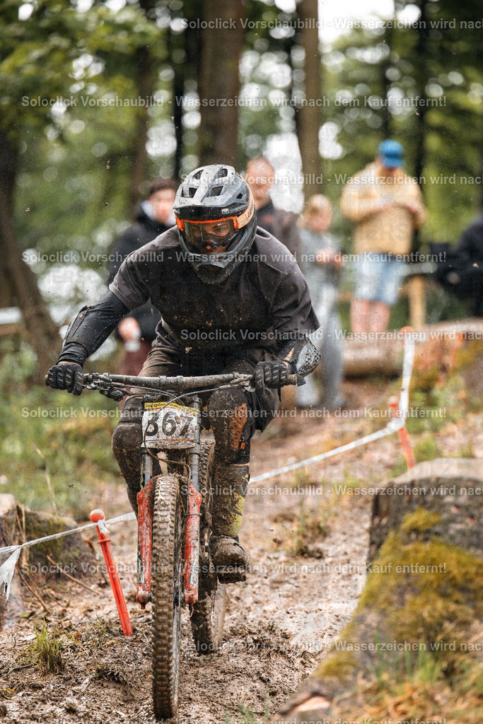Mountainbike-Action im Wald | Ein Mountainbiker in voller Ausrüstung rast auf einem schlammigen Weg durch einen dichten Wald. Er trägt einen schwarzen Helm mit einer orangefarbenen Markierung und eine dunkle Schutzkleidung. Das Fahrrad ist ebenfalls mit Schlamm bedeckt und hat einen auffälligen roten Rahmen. Im Hintergrund sind weitere Personen und Bäume zu sehen. - Realisiert mit Pictrs.com
