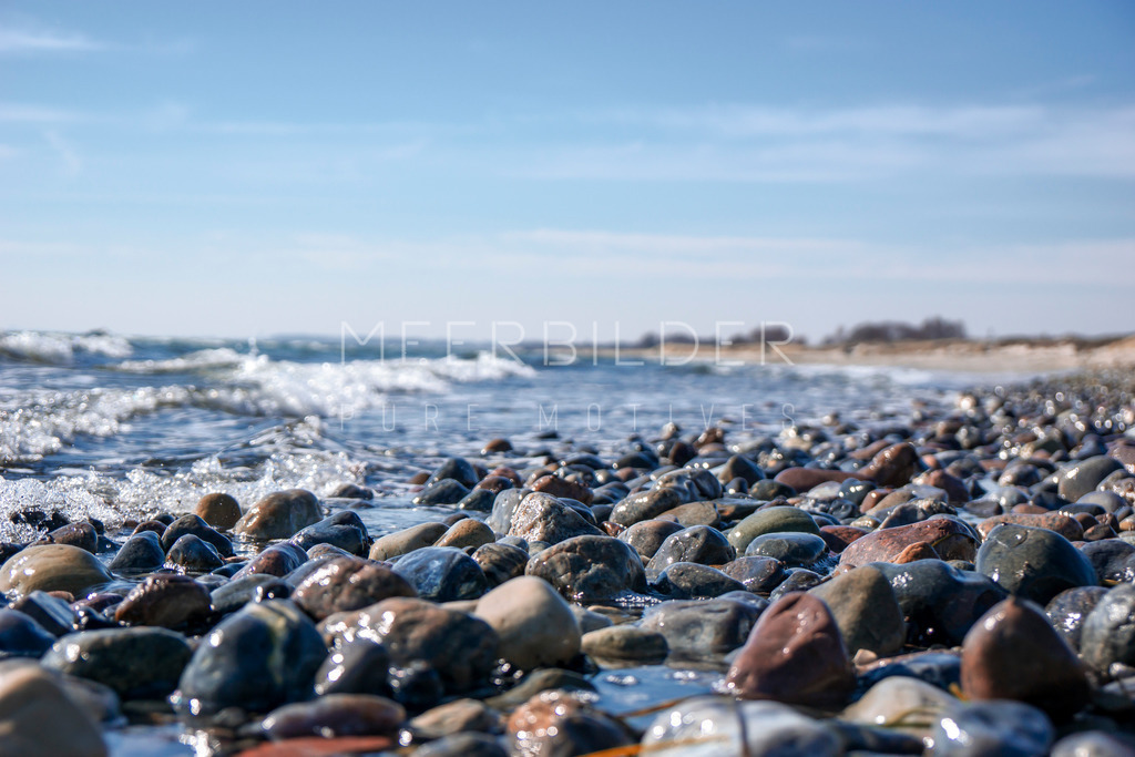 Steine und Spülsaum // Steinstrand | Steine am Spülsaum - leichte Wellen halten die Steine feucht und die Sonne bringt diese zum Glänzen. Bei jedem Auf- und Ablaufen der Wellen hört man das sanfte Grollen der Steine. Der Fokus bei diesem Motiv wurde bewusst auf die untere Mitte gesetzt, hierdurch sind die unteren Steine und das Meer im Hintergrund mit einem feinen Bokeh-Effekt versehen (Unschärfe).