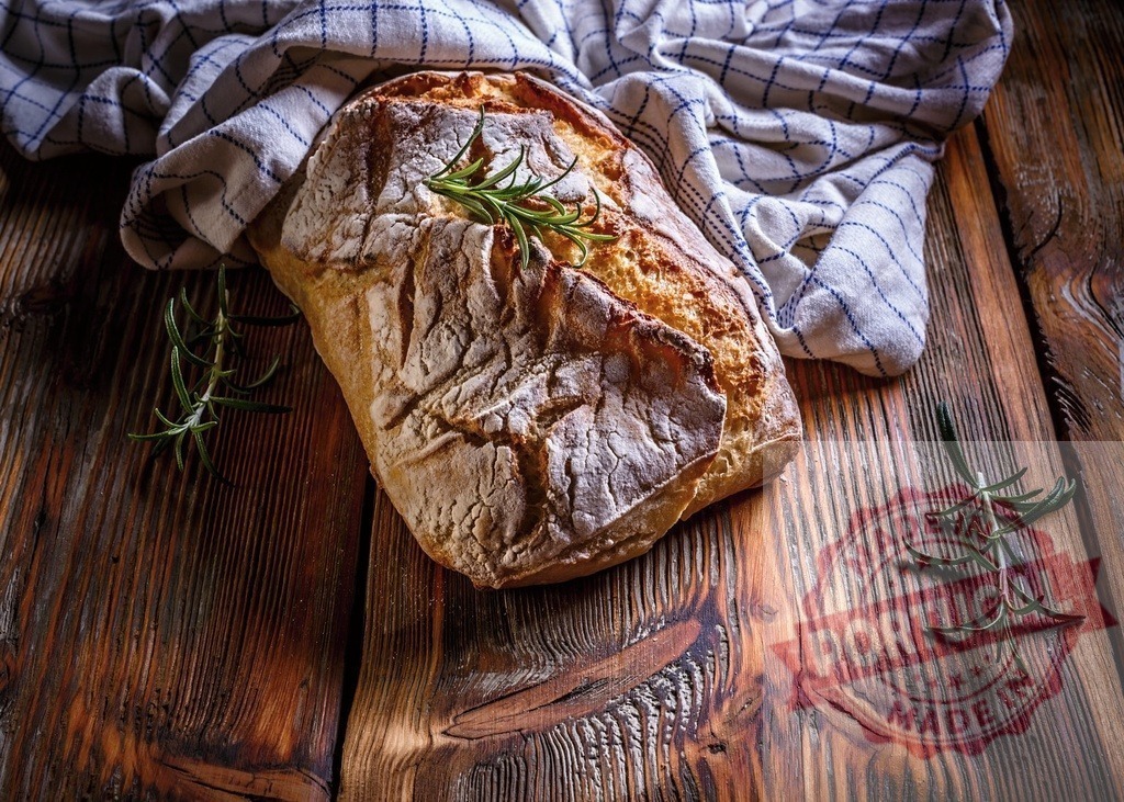 Freshly baked rustic bread with rosemary on wood | Rustic homemade bread loaf with rosemary sprig on a wooden table with a kitchen towel - Realisiert mit Pictrs.com