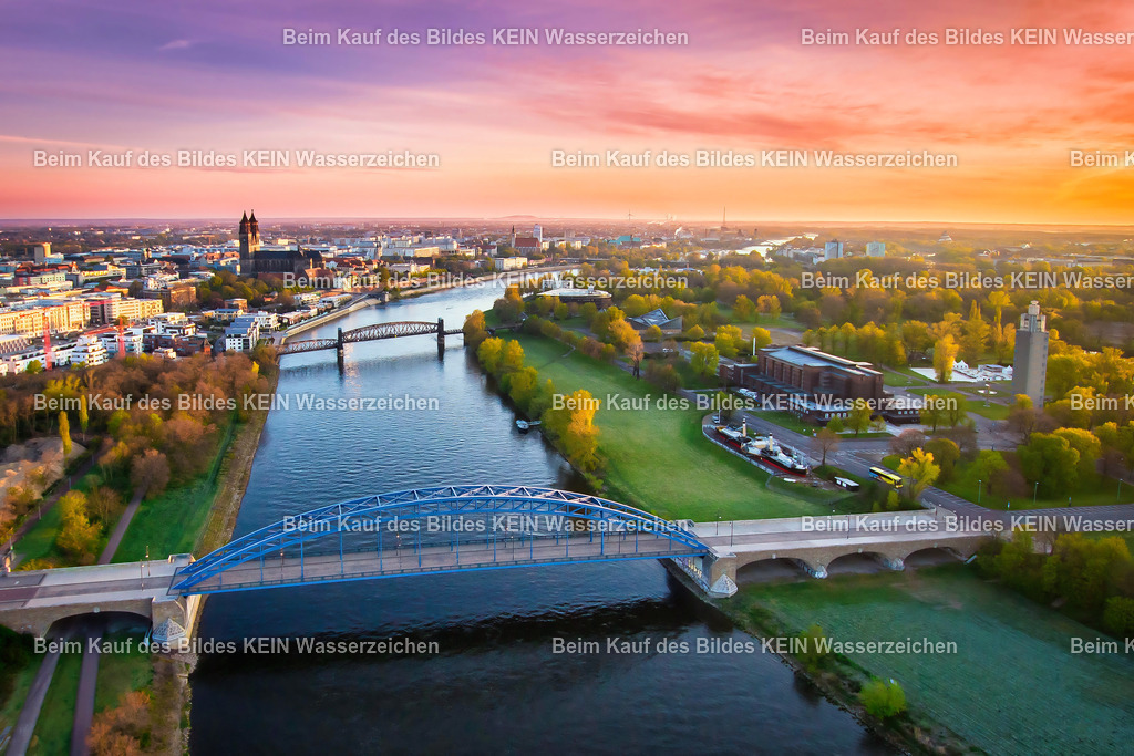 Sternbrücke im Morgenlicht | Dieses Luftbild zeigt die Sternbrücke in Magdeburg während eines eindrucksvollen Sonnenaufgangs über der Elbe. Die ersten Sonnenstrahlen des Tages tauchen den Himmel in intensive Rot-, Orange- und Magentatöne. Durch die gezielte Bildbearbeitung wird diese außergewöhnliche Lichtstimmung zusätzlich verstärkt und verleiht der Aufnahme eine besonders atmosphärische Wirkung.Im Mittelpunkt des Bildes steht die markante Stahlkonstruktion der Sternbrücke, die sich elegant über die Elbe spannt. Aus der Vogelperspektive wird ihre geschwungene Form besonders deutlich sichtbar. Die Elbe prägt als zentrales Landschaftselement die gesamte Komposition und führt den Blick durch das Bild.Eingebettet in die Flusslandschaft präsentiert sich auch der Stadtpark Rotehorn, dessen weitläufige Grünflächen einen reizvollen Kontrast zur technischen Architektur der Brücke bilden. Die Kombination aus Wasser, Parklandschaft und Ingenieurskunst macht diesen Bereich zu einem der schönsten Stadtpanoramen Magdeburgs.Dieses Luftbild verbindet die besondere Stimmung eines farbenprächtigen Morgenhimmels mit der markanten Architektur der Sternbrücke und zeigt die Elbestadt aus einer außergewöhnlichen Perspektive. - Realisiert mit Pictrs.com
