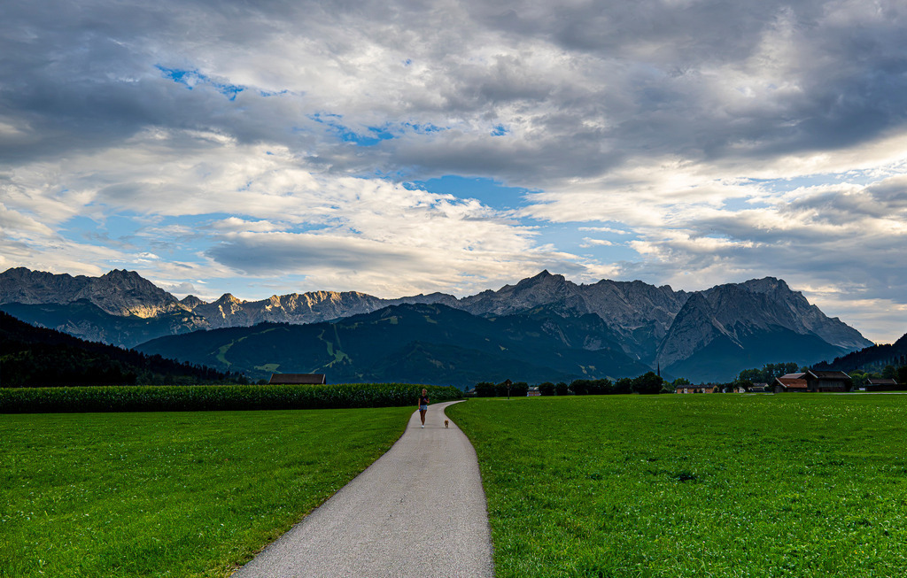 Weg ins Weite: Abendstille vor dem Zugspitzmassiv | Ein asphaltierter Feldweg schlängelt sich bei Farchant durch saftig grüne Wiesen direkt auf das imposante Zugspitzmassiv zu. In der friedlichen Weite geht eine Person mit ihrem Hund spazieren, während die feine Abendsonne die markanten Felsgipfel im Hintergrund sanft erhellt. Ein ausdrucksstarker Wolkenhimmel verleiht der Szenerie eine kraftvolle, fast mystische Atmosphäre. Das Foto strahlt tiefste Ruhe, Verbundenheit mit der Natur und den Zauber eines idyllischen Feierabends in den Alpen aus. - Realisiert mit Pictrs.com
