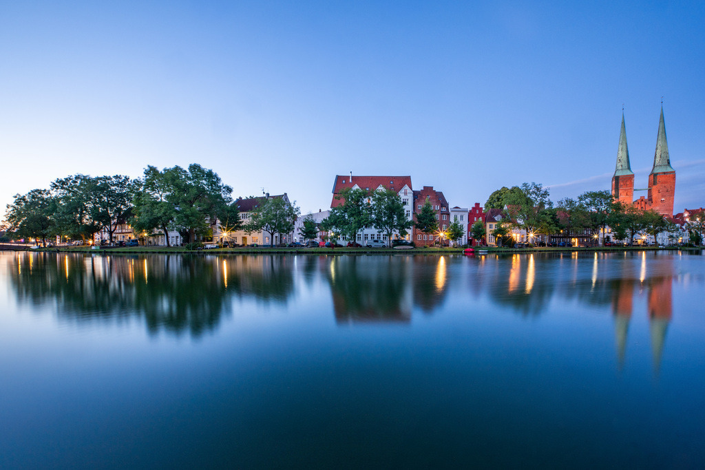 Lübeck am Wasser in der blauen Stunde | Die Lübecker Stadtsilhouette am ruhigen Wasser in der blauen Stunde, mit klaren Spiegelungen und den markanten Türmen der Altstadt. - Realisiert mit Pictrs.com