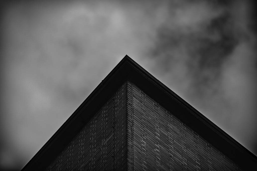 Minimalist Architecture – Urban Geometry in Black and White | This black and white photograph captures a minimalist architectural perspective from a low angle. The sharp lines of the building lead the viewer’s eye upward into a dramatic sky, creating a strong geometric composition.The reduced visual language of form, structure, and light gives this image a modern fine art character. The brick texture and symmetrical perspective emphasize the graphic impact of the architecture.This photograph works perfectly as a high-quality wall art print for modern interiors, offices, or architectural spaces. - Realisiert mit Pictrs.com
