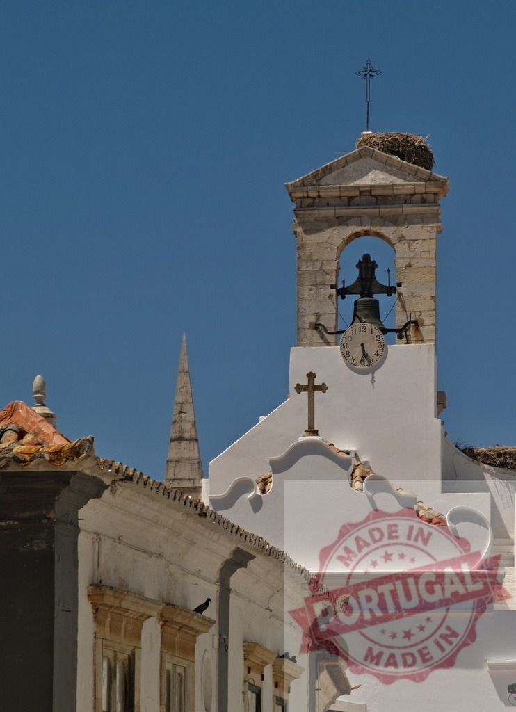 Faro church bell tower with stork nest and clock | Faro church bell tower featuring a stork nest, clock, and cross against a blue sky - Realized with Pictrs.com