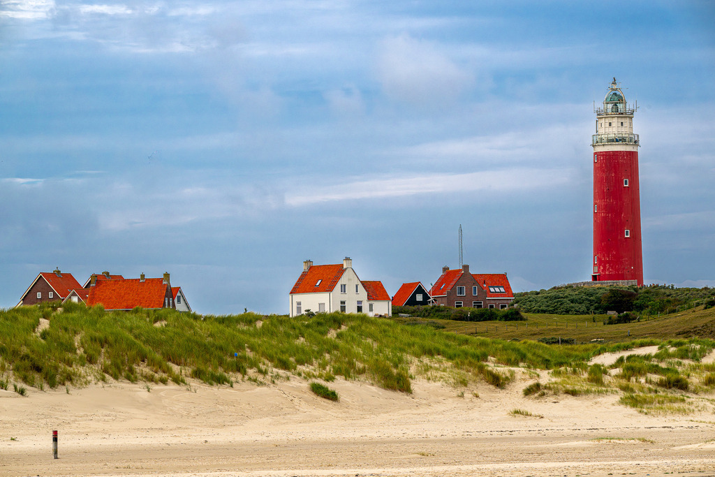 Maritimes Wahrzeichen von Texel: Leuchtturm Eierland und Küstenhäuser hinter den Dünen bei De Cocksdorp | Der leuchtend rote Leuchtturm Eierland überragt die Dünenlandschaft und typischen Wohnhäuser mit roten Ziegeldächern am Nordseestrand von Texel bei De Cocksdorp. Im Vordergrund erstreckt sich ein breiter Sandstrand mit bewachsenen Dünenketten unter leicht bewölktem Himmel. Das Motiv eignet sich ideal für redaktionelle Beiträge über Tourismus auf den Westfriesischen Inseln, Seefahrt, Ausflugsziele in den Niederlanden und maritime Küstenarchitektur. - Realisiert mit Pictrs.com