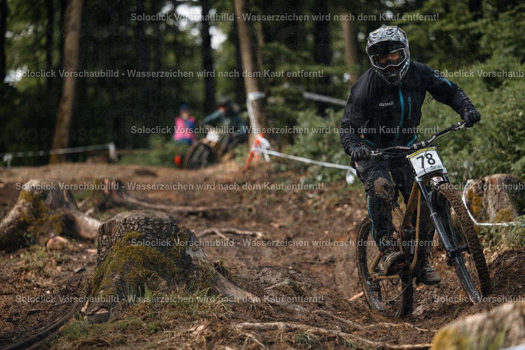 Mountainbike-Action im Wald | Ein Mountainbiker in voller Ausrüstung rast einen anspruchsvollen Trail hinunter. Der Fahrer trägt einen schwarzen Helm mit einer weißen Visier, eine schwarze Jacke und Handschuhe. Er trägt die Nummer 78 auf seinem Rücken. Der Trail ist staubig und mit Wurzeln und Steinen übersät. Im Hintergrund sind weitere Mountainbiker und Bäume zu sehen. - Realisiert mit Pictrs.com