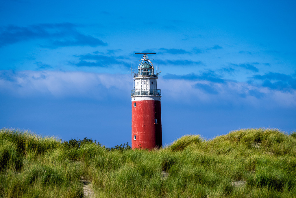 Rot-weißer Leuchtturm Eierland in den Dünen auf der Nordseeinsel Texel | Der charakteristische rote Leuchtturm Eierland ragt an der Nordspitze der niederländischen Insel Texel über die saftig grünen Dünen hinaus. Die Aufnahme zeigt das markante Wahrzeichen bei leicht bewölktem, blauem Himmel. Das Motiv eignet sich ideal für redaktionelle Berichte über den Tourismus in den Niederlanden, Urlaub an der Nordsee, maritime Architektur, Küstenschutz sowie Reiseberichte über die Westfriesischen Inseln. - Realisiert mit Pictrs.com
