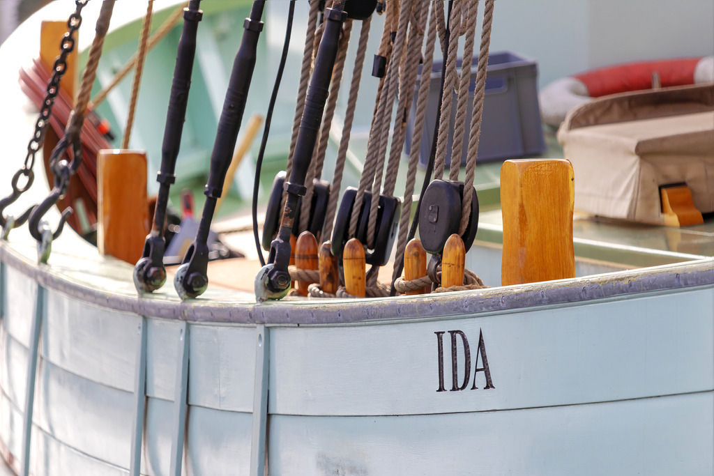 Wandbild: Boot in Nahaufnahme | Maritimes Detail voller Charme – dieses Wandbild zeigt eine Nahaufnahme eines Bootes mit seinem Namen „IDA“, eingerahmt von Seilen und Rollen aus der Takelage. Die feine handwerkliche Verarbeitung und die warmen Farbtöne der Bootselemente verleihen der Szene eine nostalgische und maritime Ausstrahlung. Ein ideales Kunstwerk für alle, die die Liebe zum Segeln und die Schönheit traditioneller Bootskonstruktionen in ihr Zuhause bringen möchten. - Realisiert mit Pictrs.com