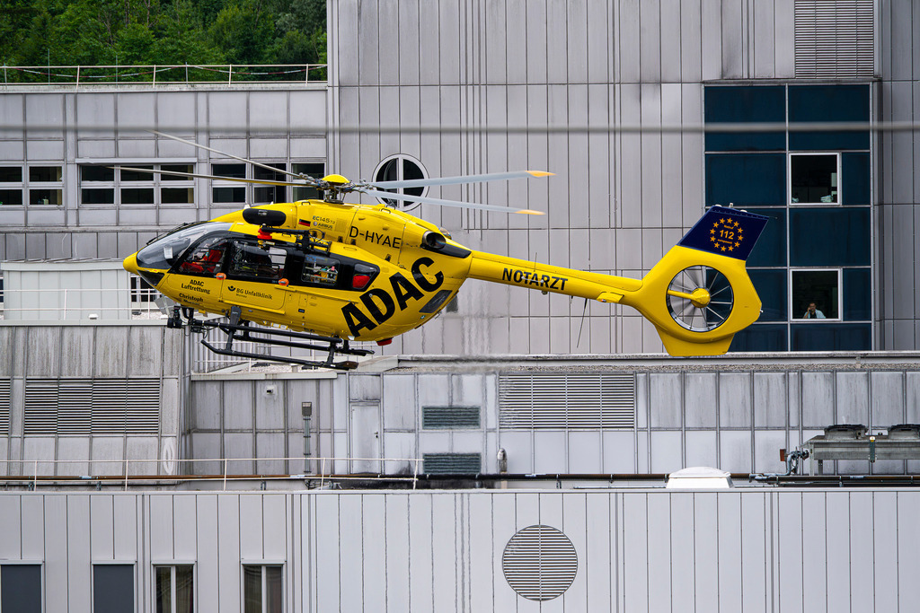 Hoffnung im Anflug: ADAC-Rettungshubschrauber am Klinikum Garmisch-Partenkirchen | Ein leuchtend gelber ADAC-Rettungshubschrauber der Type H145 schwebt im Präzisionsflug direkt vor der modernen Fassade des Klinikums Garmisch-Partenkirchen. Das Motiv fängt die Anspannung und Professionalität eines medizinischen Notfalleinsatzes ein. Das Zusammenspiel aus modernster Technik, schneller Hilfe und der markanten Architektur der Klinik vermittelt ein tiefes Gefühl von Sicherheit, Verlässlichkeit und Hoffnung in kritischen Momenten – wenn jede Sekunde zählt, um Menschenleben zu retten. - Realisiert mit Pictrs.com