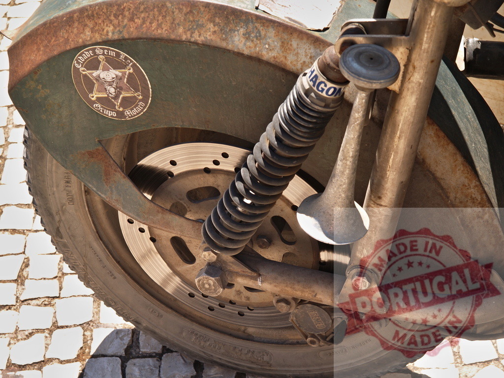 Vintage custom motorcycle wheel showing rust and shock absorber | Old motorcycle wheel with fender, rustic metal, tire, brake disk, and shock absorber on pavement - Realized with Pictrs.com
