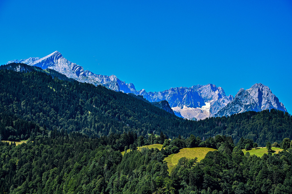 Gipfelglück über Garmisch-Partenkirchen | Dieses atemberaubende Panorama fängt die unberührte Schönheit des Werdenfelser Landes ein. Unter einem tiefblauen Himmel erstreckt sich die majestätische Bergkulisse von der markanten Alpspitze bis hin zur mächtigen Zugspitze. Dichte, dunkelgrüne Bergwälder bedecken die Hänge im Vordergrund, durchbrochen von saftigen, sonnenbeschienenen Almwiesen nahe Garmisch-Partenkirchen. Die Szene strahlt eine erhabene Ruhe und die zeitlose Kraft der bayerischen Hochalpen aus – ein Bild wie aus dem Bilderbuch. - Realisiert mit Pictrs.com
