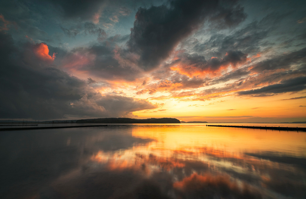 Abendfeuer – Sonnenuntergang über dem Jasmunder Bodden | Der Himmel brennt in warmen Farben, während die untergehende Sonne die Landschaft auf Rügen in ein goldenes Licht taucht. Als Panorama betont das Bild die weite Stimmung des Augenblicks. - Realisiert mit Pictrs.com