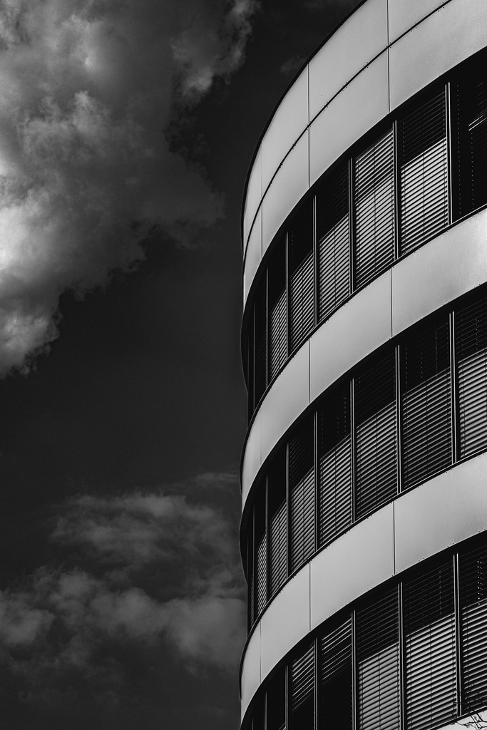 Curves of Contrast | Sanft geschwungene Architektur trifft auf einen dramatischen Himmel in einer kontrastreichen Schwarzweißkomposition. Die fließenden Linien der modernen Fassade stehen im spannenden Gegensatz zu den kraftvollen Wolkenformationen und erzeugen eine harmonische Verbindung aus Eleganz, Geometrie und Atmosphäre. Die reduzierte Bildsprache lenkt den Blick auf Form, Licht und Struktur und macht dieses Motiv zu einem zeitlosen Fine-Art-Print für moderne Räume. - Realisiert mit Pictrs.com