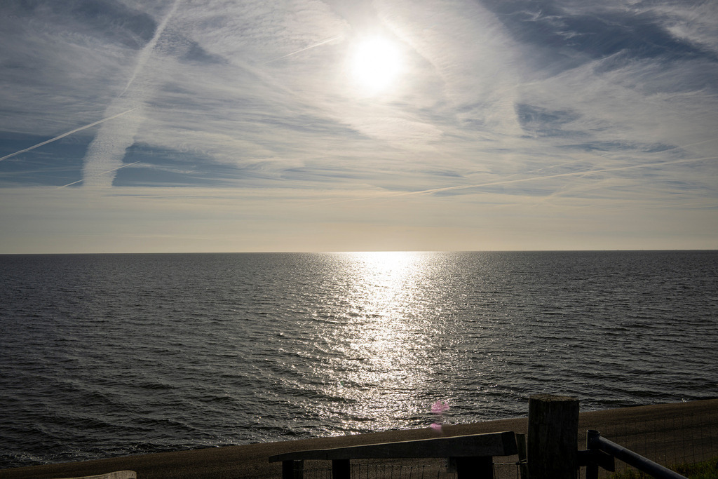 Sonnenreflexion auf der Nordsee vor der Insel Texel: Weite Küstenlandschaft am Deich | Blick von einer Deichüberquerung über einen Küstenweg auf die ruhige Nordsee bei Texel. Die hell im Dunst stehende Sonne spiegelt sich mit einer gleißenden Lichtspur im leicht welligen Wasser. Ein leicht bewölkter Himmel mit sichtbaren Kondensstreifen erstreckt sich über den weiten Horizont der westfriesischen Insel. Das Motiv fängt die friedliche, kühle Atmosphäre der niederländischen Nordseeküste ein - Realisiert mit Pictrs.com