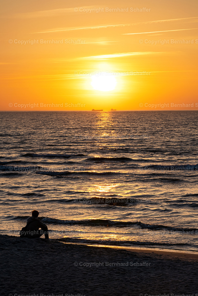 Sunset am Meer | Intensiver Sonnenuntergang am Sandstrand der Ostsee in Warnemünde in Deutschland. Eine Person sitzt im Sand und blickt sehnsüchtig auf zwei ferne Schiffe am Horizont. - Realisiert mit Pictrs.com