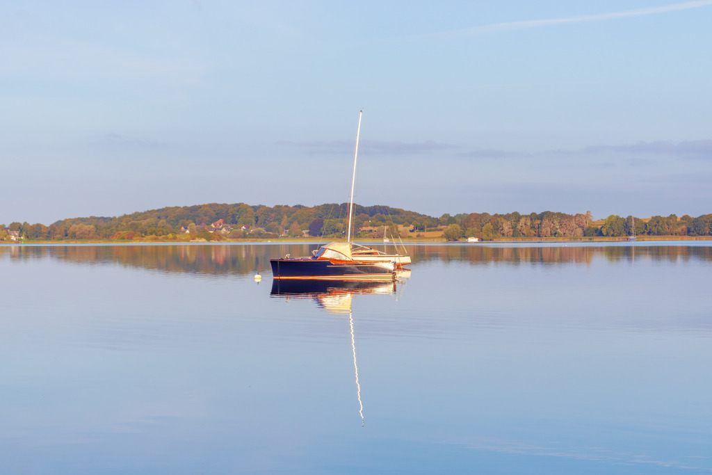 Wandbild: Segelboote spiegelt sich auf der Schlei | Dieses Wandbild im Querformat zeigt ein Segelboot in Sieseby das sich auf dem ruhigen Wasser der Schlei spiegelt. In der Ferne kann man auf der anderen Uferseite der Schlei einen Wald sehen. Dieser Spiegelt sich auch auf der Wasseroberfläche.  - Realisiert mit Pictrs.com