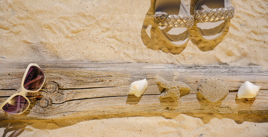golden sun shines at elements on the beach sand D | Goldenes Sonnenlicht scheint auf einen Sandstrand. Goldene Damensandalen, eine Sonnenbrille, Treibholz, Muscheln und Sandfiguren bieten einen sommerlichen Hinterund mit Copy Space. Thema Urlaub. 