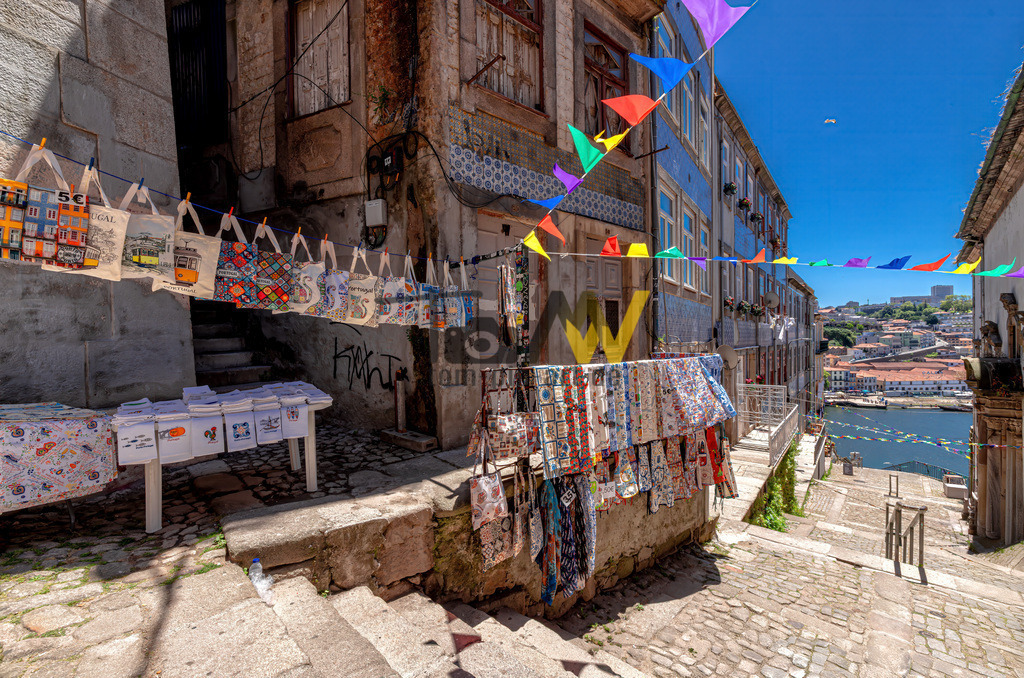 Eine Gasse in Porto, bunt dekoriert,blauer Himmel,Meerblick | Das Bild zeigt eine Szene aus der historischen Altstadt von Porto, Portugal. Die Szene zeigt eine steile, gepflasterte Straße oder Treppe, die hinunter in Richtung des Douro-Flusses führt.An der linken Seite sind Verkaufsstände mit lokalen Textilien und Souvenirs aufgebaut.Bunte Fähnchen sind über die Gasse gespannt und sorgen für eine festliche Atmosphäre. - Realisiert mit Pictrs.com