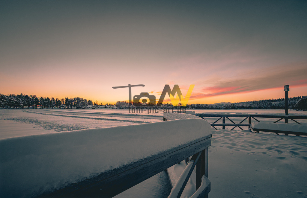 Verschneiter Holzsteg bei magischem Sonnenaufgang---Winter | Das Bild zeigt eine Winterlandschaft bei Sonnenaufgang in Schweden. Eine verschneite Holzbrücke oder ein Steg führt in die Ferne.Die Sonne taucht den Horizont in warme Orange- und Rosatöne.Die umliegende Landschaft ist von tiefem Schnee bedeckt.Am Horizont sind dunkle Baumreihen (Nadelwälder) zu sehen. - Realisiert mit Pictrs.com
