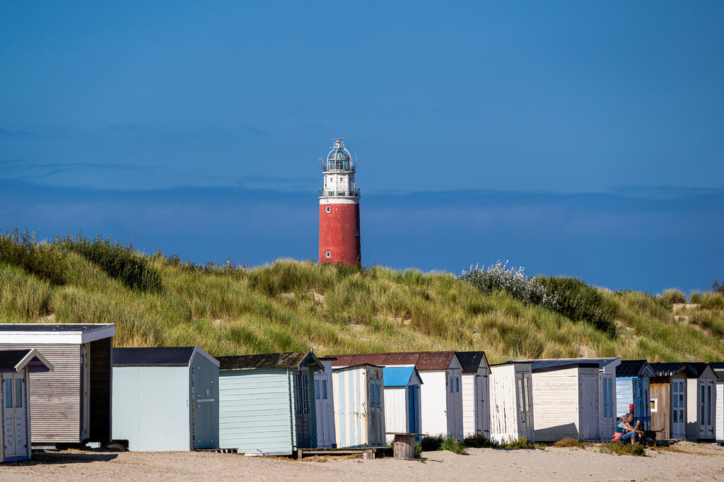 Wahrzeichen von Texel: Roter Leuchtturm hinter Strandhäuschen und Dünen in De Cocksdorp | Der markante rote Leuchtturm Eierland ragt an der Nordspitze der Insel Texel hinter bewachsenen Dünen empor. Im Vordergrund beruhen charakteristische, bunt gestrichene Strandhäuschen auf dem Sandstrand vor strahlend blauem Himmel. Das stimmungsvolle Motiv vereint typische Elemente der niederländischen Küstenlandschaft und eignet sich ideal für Themen rund um Tourismus, Urlaub an der Nordsee, Reiseziele und maritime Architektur. - Realisiert mit Pictrs.com
