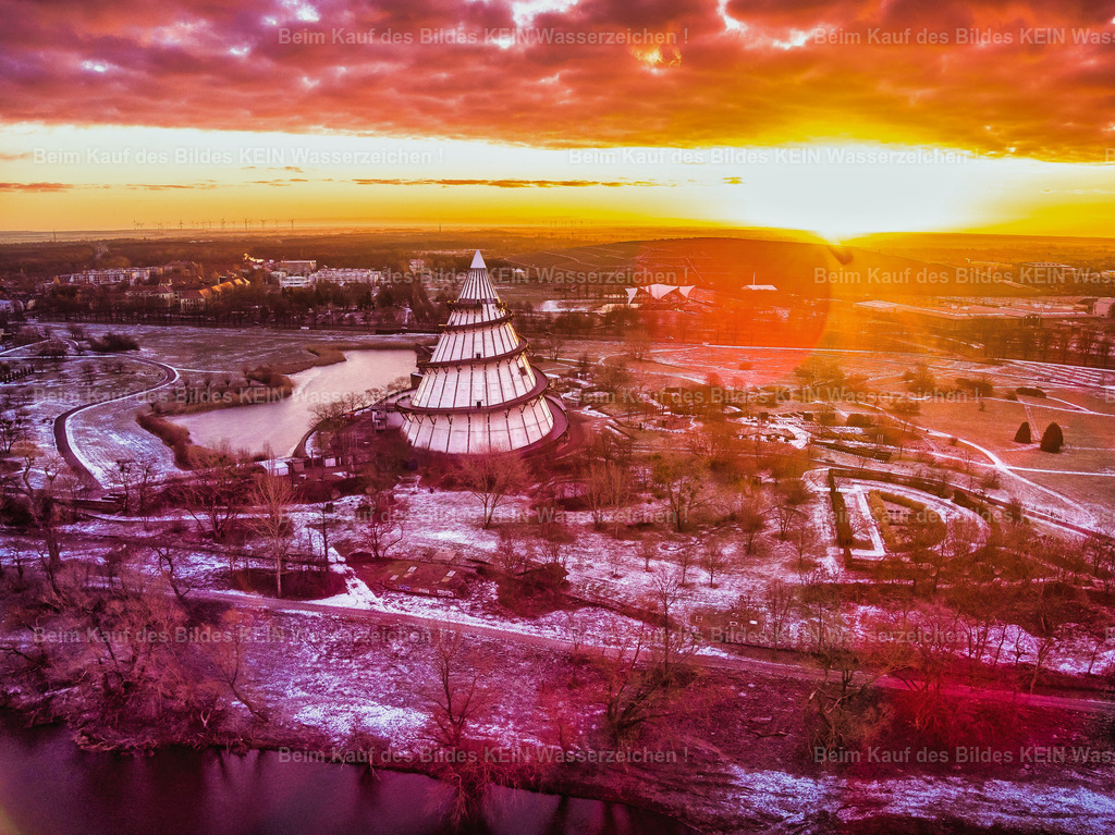 Magdeburg_Luftbild_Jahrtausendturm_im_roten_Morgenlicht_Sonnenaufgang_LR-0034 | Dieses Luftbild zeigt den Jahrtausendturm im Magdeburger Elbauenpark während eines winterlichen Sonnenaufgangs. Die Aufnahme entstand in den frühen Morgenstunden, als die aufgehende Sonne den Himmel in intensive Rot- und Orangetöne tauchte. Durch die Gegenlichtsituation und die gezielte Bildbearbeitung wird die besondere Atmosphäre dieses Moments zusätzlich betont.Der Jahrtausendturm, eines der bekanntesten Wahrzeichen Magdeburgs, hebt sich eindrucksvoll vor dem farbenprächtigen Himmel ab. Die kahlen Bäume des Winters und die leicht mit Schnee bedeckten Flächen verleihen der Szene eine ruhige und fast märchenhafte Stimmung. Aus der Luft wird die einzigartige Lage des Turms im weitläufigen Elbauenpark besonders deutlich.Der Jahrtausendturm gilt mit seinen 60 Metern Höhe als einer der höchsten Holztürme seiner Art weltweit und prägt die Silhouette des Parks seit der Bundesgartenschau 1999. Das Zusammenspiel aus moderner Architektur, winterlicher Landschaft und spektakulärem Morgenlicht macht diese Luftaufnahme zu einem besonderen Blick auf die Landeshauptstadt Magdeburg. - Realisiert mit Pictrs.com