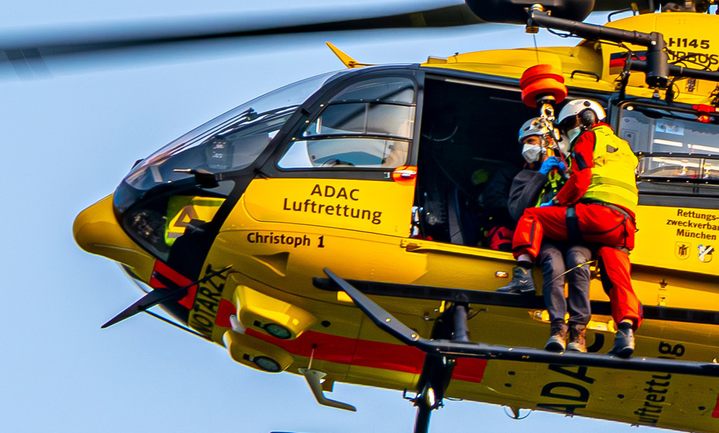 Bergretter | Ein markanter gelber ADAC-Rettungshubschrauber (Christoph 1) befindet sich im Schwebeflug vor einem klaren, hellen Himmel. An der Seite ist die Rettungswinde im Einsatz: Ein Luftretter sichert eine Person, während sie gemeinsam am Seil unterhalb der offenen Kabinentür hängen – ein intensiver Einblick in die lebensrettende Arbeit der Bergwacht oder Luftrettung. - Realisiert mit Pictrs.com