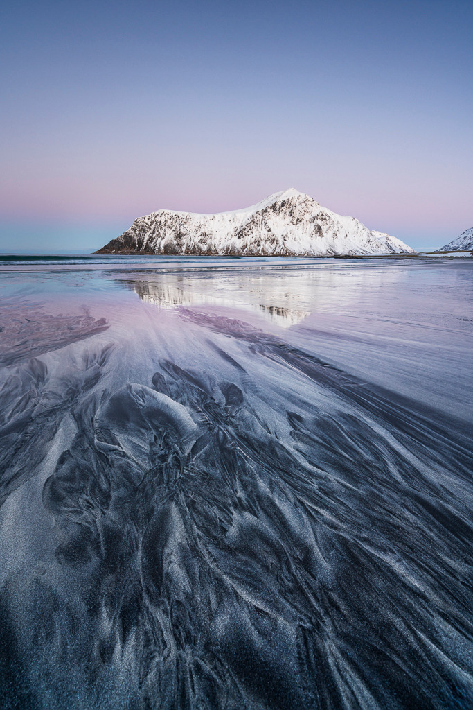Strukturen im Sand – Flakstad Beach, Lofoten | Am Flakstad Beach auf den Lofoten treffen grafische Sandstrukturen auf das sanfte Licht des Nordens. Der verschneite Berg spiegelt sich im ruhigen Wasser – ein Bild, das Minimalismus, Ruhe und natürliche Textur vereint. - Realisiert mit Pictrs.com
