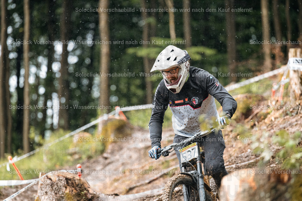 Mountainbiker im Wald | Ein Mountainbiker rast einen staubigen, schmalen Trail in einem bewaldeten Gebiet hinunter. Er trägt einen weißen Helm mit einer schwarzen Visier, eine Schutzbrille und eine schwarze Jacke mit weißen Akzenten. Er trägt auch Handschuhe und eine Nummer 37 ist auf seinem Trikot zu sehen. Der Trail ist mit Baumstümpfen und Wurzeln übersät, und der Wald ist dicht und grün. Es regnet leicht. - Realisiert mit Pictrs.com