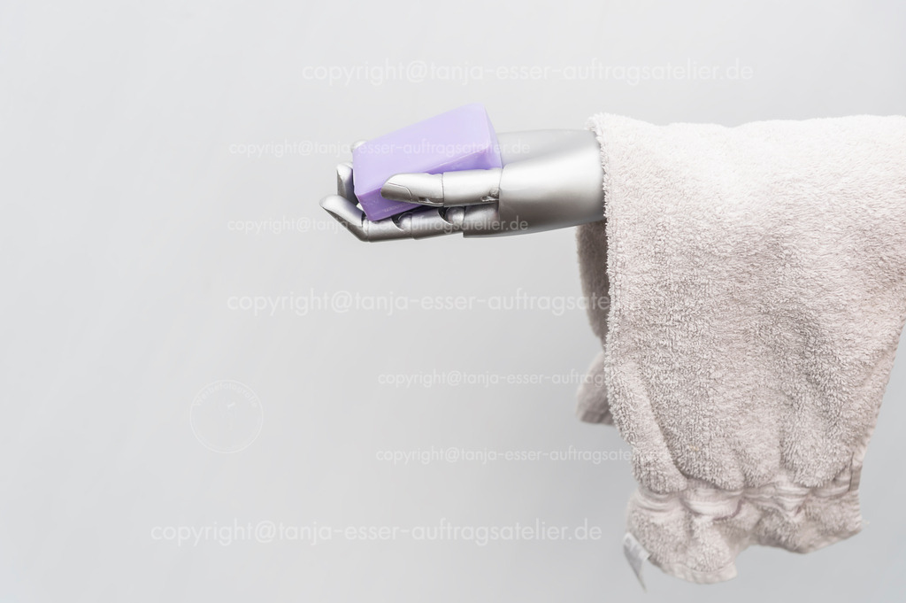 Robot takes care of people | Der Roboterarm hält Frotteehandtücher und ein Stück Seife in der Hand. Das Bild symbolisiert die Pflege von Menschen durch Maschinen. Konzeptuelles Foto vor weißem Hintergrund mit Copy Space.