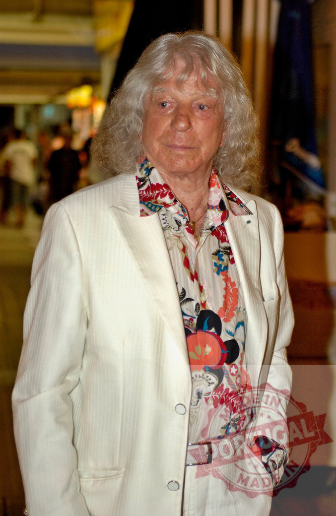 Elegant senior man wearing white suit and floral shirt posing indoors | Stylish older man with long gray hair and white suit standing indoors, looking confident and fashionable - Realisiert mit Pictrs.com