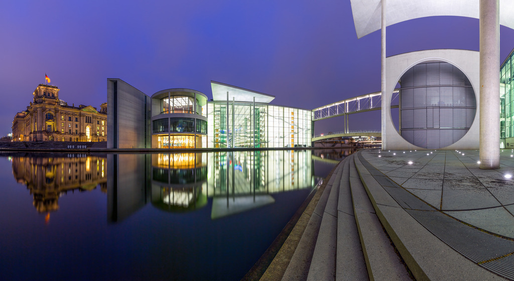 Wandbild - Berlin bei Nacht: Architektur der Vergangenheit und Zukunft | Dieses eindrucksvolle Panorama zeigt die nächtliche Szenerie des Regierungsviertels in Berlin. Links erstrahlt der historische Reichstag majestätisch im warmen Licht, während die modernen Gebäude des Marie-Elisabeth-Lüders-Hauses und des Paul-Löbe-Hauses rechts mit ihrer gläsernen Architektur und klaren Linien beeindrucken. Die Spiegelungen der Gebäude im ruhigen Wasser der Spree verstärken die Schönheit dieser Komposition. Die Verbindung von Alt und Neu, Historie und Moderne, verleiht diesem Bild eine besondere Tiefe und zeigt Berlin als eine Stadt, die ihre Vergangenheit ehrt und gleichzeitig zukunftsorientiert ist.