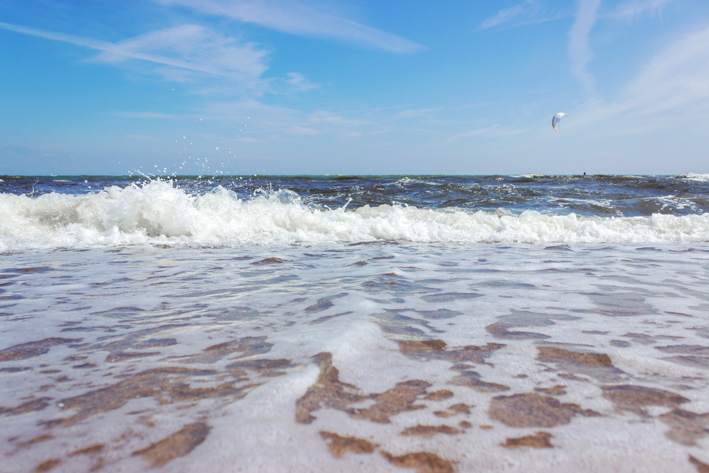 Wandbild: Dynamische Küstenschönheit in Weidefeld | Dieses beeindruckende Wandbild im Querformat zeigt eine auslaufende Welle am Strand in Weidefeld. Das klare Wasser ermöglicht einen Blick auf den darunterliegenden Sandstrand. In der Ferne zieht ein Kitesurfer seine Bahnen, was dem Bild eine dynamische Note verleiht. Der fast wolkenlose, blaue Himmel rundet die Szenerie ab und schafft eine ruhige, maritime Atmosphäre. - Realisiert mit Pictrs.com