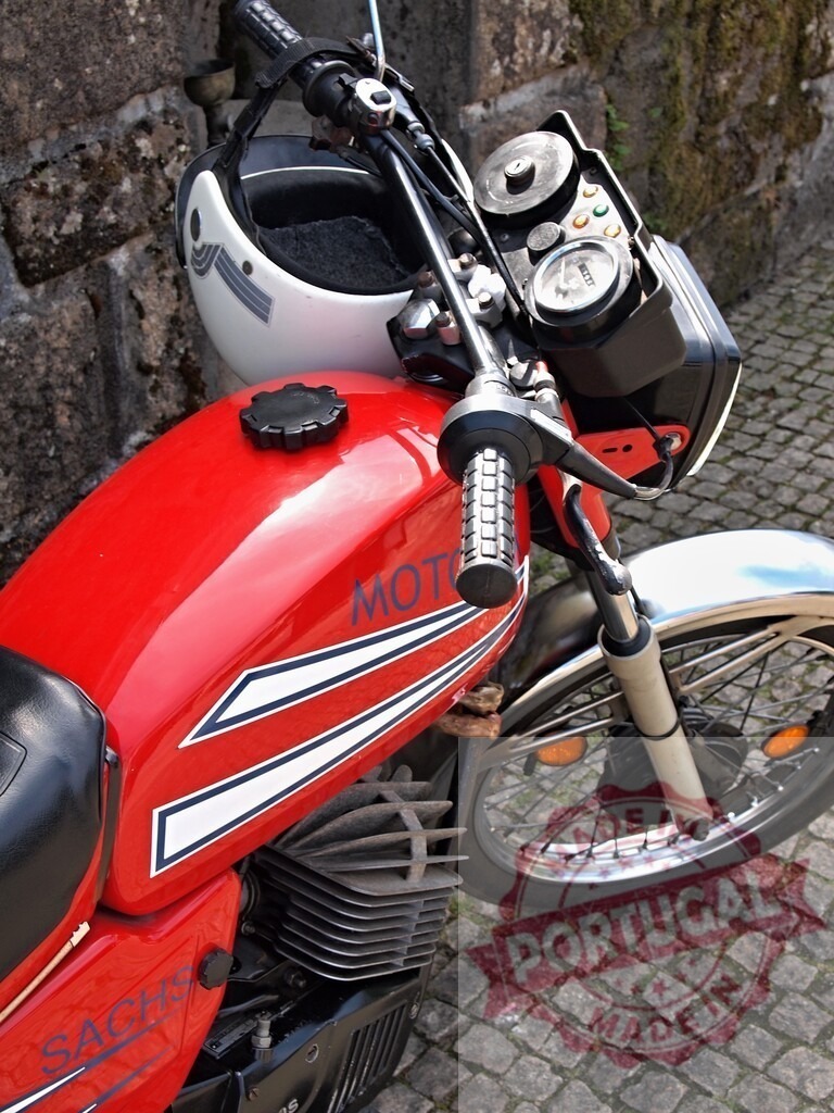 Red Sachs Motorcycle Parked with Helmet on Cobblestone Street | Red Sachs motorcycle parked on a cobblestone street with a white helmet hanging from the handlebars - Realized with Pictrs.com