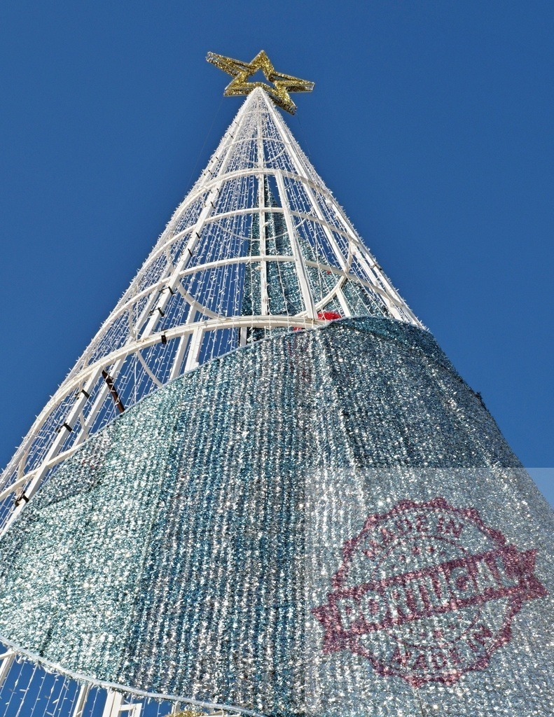 Giant christmas tree with sparkling blue decorations and golden star against blue sky | Modern giant christmas tree with sparkling blue decorations and golden star against blue sky celebrating christmas time - Realized with Pictrs.com