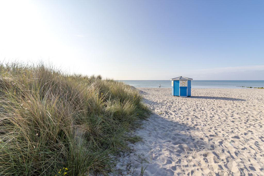 Wandbild: Zwischen Küstengras und Horizont – Ruhe an der Ostsee | Dieses Wandbild zeigt eine ruhige Strandszene in Grömitz an einem klaren Morgen. Der helle Sand zieht sich sanft bis zum Horizont, wo die Ostsee ruhig und weit unter einem fast wolkenlosen Himmel liegt. Links im Bild wächst hohes Küstengras auf einer kleinen Erhebung und bringt Struktur und Natürlichkeit in die Komposition. Im rechten Bildbereich steht ein kleines blau-weißes Strandhäuschen. Die Szene wirkt geordnet und zugleich entspannt, das Licht fällt weich über die gesamte Fläche und unterstreicht die friedliche Atmosphäre. Dieses Motiv eignet sich ideal als Wandbild für maritime Wohnkonzepte – ob als Leinwandbild, Acrylglasbild, Alu-Dibond FineArt Print oder als Akustikbild. Ein stilvoller Akzent für Wohnzimmer, Büro oder Ferienwohnung. - Realisiert mit Pictrs.com