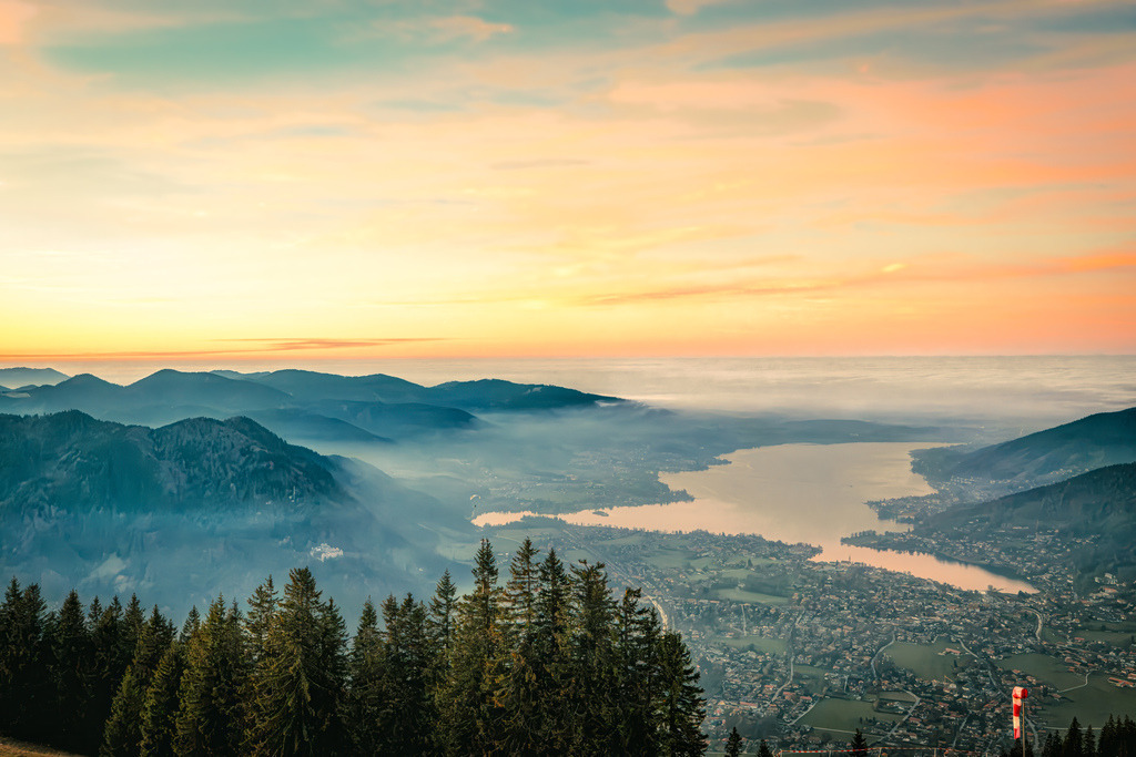 Malerische Aussicht auf der mit leichtem Dunst überzogenen Tegernsee bei Sonnenuntergang | Das ausgewählte Foto zeigt eine malerische Aussicht auf den Tegernsee in den bayerischen Voralpen, aufgenommen bei einem stimmungsvollen Sonnenuntergang.Diese charakteristische Perspektive – mit dem Blick hinab auf den langgezogenen See, die umliegenden Ortschaften wie Rottach-Egern und Tegernsee sowie die sanften Hügelketten im Hintergrund – wird typischerweise vom Wallberg aus fotografiert - Realisiert mit Pictrs.com