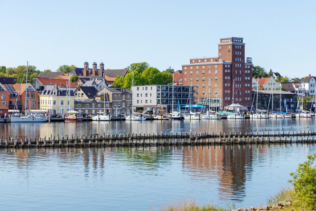Wandbild: Spiegelung am Hafen in Kappeln an der Schlei | Dieses Wandbild im Querformat zeigt den Hafen in Kappeln an der Schlei. Auf dem ruhigen Wasser der Schlei spiegeln sich die Gebäude am Hafen. Die Spiegelung wird nur von dem Heringszaun inmitten der Schlei unterbrochen.  - Realisiert mit Pictrs.com