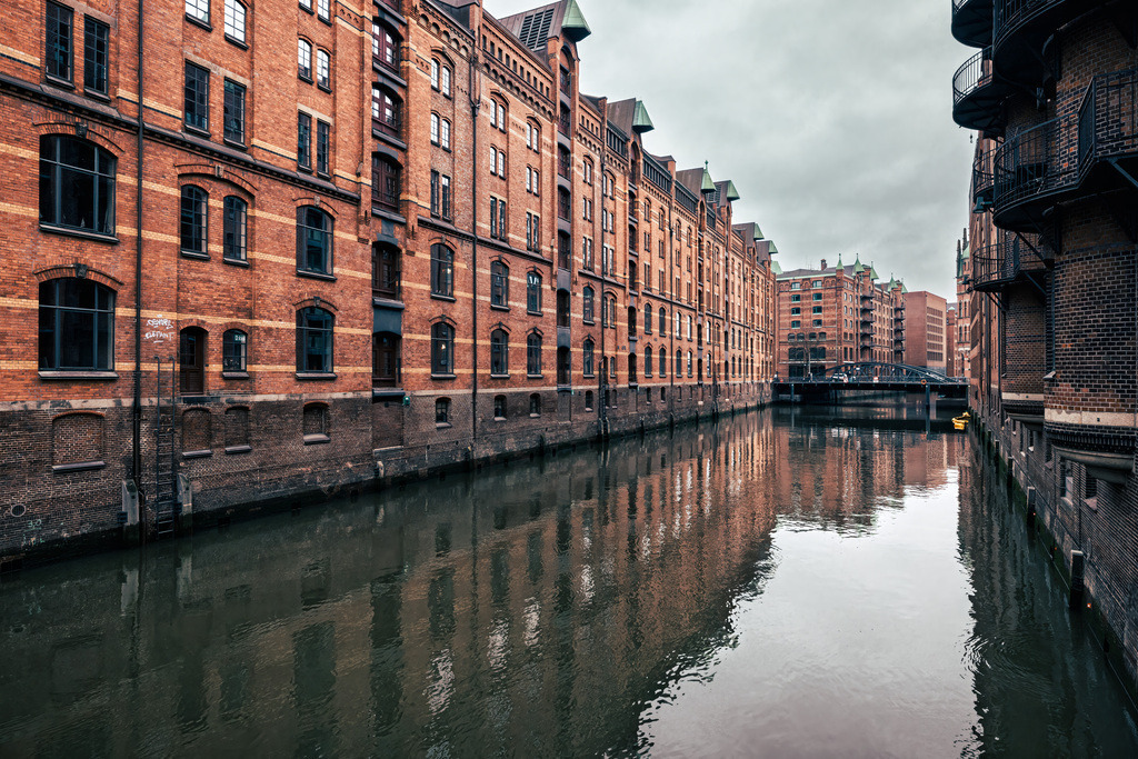 Lagerkomplex der Speicherstadt Hamburg spiegelt sich im Wassser | Das Bild zeigt die Hamburger Speicherstadt. Es handelt sich um den größten zusammenhängenden historischen Lagerhauskomplex der Welt, der zum UNESCO-Weltkulturerbe gehört. Die Speicherstadt wurde zwischen 1885 und 1927 erbaut. Sie liegt südlich der Hamburger Altstadt, zwischen dem Oberhafen und dem Sandtorkai. Die charakteristische Architektur aus roten Backsteingebäuden und Fleeten (Kanälen) spiegelt sich im Wasser wider. - Realisiert mit Pictrs.com