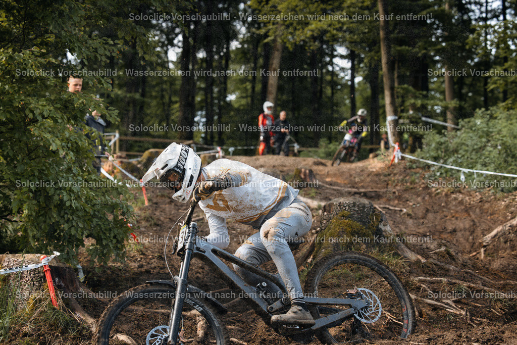 Mountainbike-Abenteuer | Ein Mountainbiker in voller Ausrüstung rast einen staubigen, holprigen Pfad in einem bewaldeten Gebiet hinunter. Er trägt einen weißen Helm und einen weißen Overall, der mit Schlamm bedeckt ist. Das Gelände ist steil und mit Wurzeln und Steinen übersät. Im Hintergrund sind weitere Personen und Bäume zu sehen. - Realisiert mit Pictrs.com