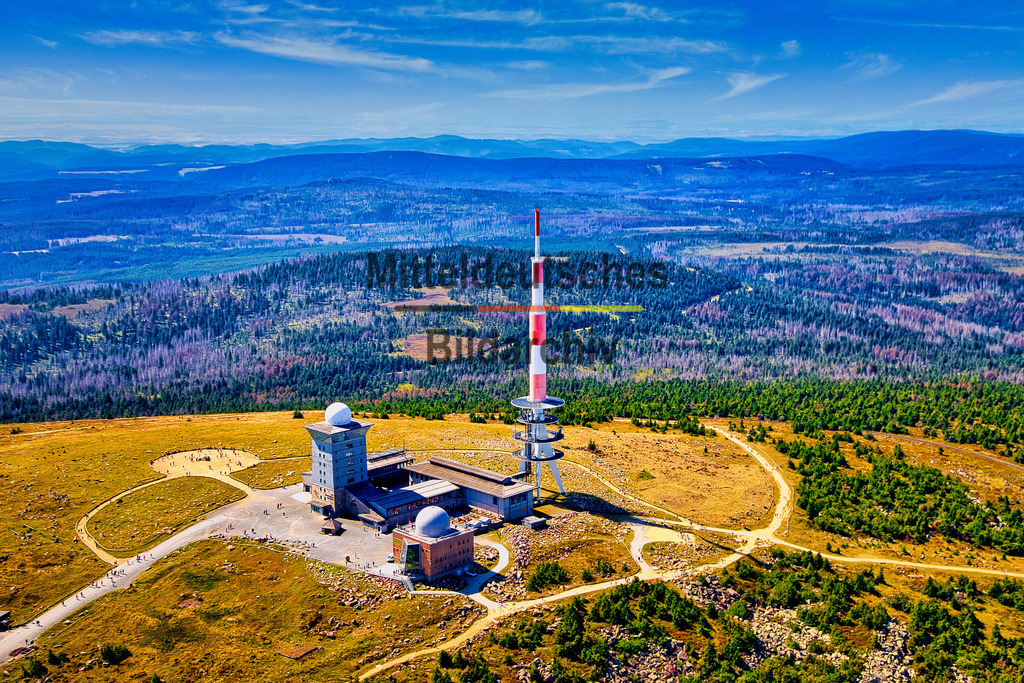 Brocken_Luftbild_Tragschrauber_Übersicht_Sendemast_8388 | Das Luftbild zeigt den Brockengipfel aus einem Tragschrauber aus geringer Entfernung. Im Mittelpunkt der Aufnahme stehen der markante Sendemast sowie die technischen und historischen Gebäude auf dem Gipfelplateau. Die nahe Vogelperspektive ermöglicht einen außergewöhnlich detaillierten Blick auf das Zusammenspiel von Funktechnik, Wetterbeobachtung und den Bauwerken auf dem höchsten Berg des Harzes.Mit 1.141 Metern über dem Meeresspiegel ist der Brocken der höchste Berg des Harzes und ganz Norddeutschlands. Das Gipfelplateau wird von der markanten Sendeanlage dominiert. Der heutige Sendeturm erreicht eine Höhe von 168 Metern und dient der Ausstrahlung von Fernseh- und Hörfunkprogrammen sowie weiteren Funkdiensten. Neben dem Sendeturm befinden sich unter anderem die Wetterstation des Deutschen Wetterdienstes, das Brockenhotel und das Brockenhaus, das Besucherzentrum des Nationalparks Harz. Die Aufnahme aus dem Tragschrauber verdeutlicht die technische Bedeutung des Brockens als herausragender Standort für Rundfunk, Wetterbeobachtung und Kommunikation. Gleichzeitig vermittelt sie einen eindrucksvollen Blick auf eines der bekanntesten Gipfelplateaus Deutschlands, auf dem moderne Technik und die außergewöhnliche Landschaft des Harzes unmittelbar aufeinandertreffen. - Realisiert mit Pictrs.com