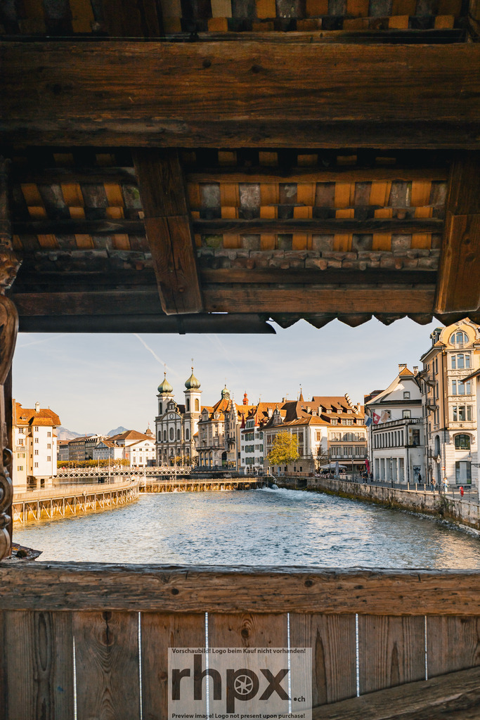 Blick durch die Spreuerbrücke auf die Luzerner Altstadt | Gebälk. Fluss. Wahrzeichen.Durch das historische Holz der Spreuerbrücke öffnet sich der Blick auf die sonnenbeschienene Uferpromenade und die barocken Türme der Jesuitenkirche. Ein einzigartiges Fenster in die Geschichte Luzerns, wo Stadt und Wasser harmonisch verschmelzen.Wähle unter "Produktauswahl" dein Wunschformat: Vom klassischen Wandbild über Puzzle &amp; Tassen bis zum digitalen Download (z.B. als Handy-Hintergrund). - Realisiert mit Pictrs.com