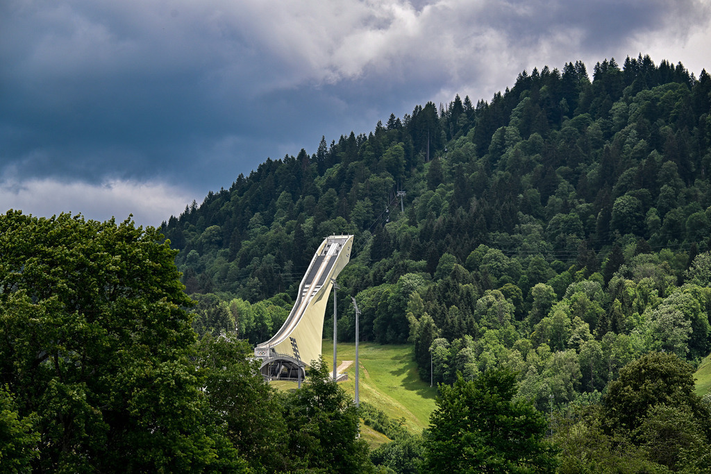 Gigant aus Stahl im grünen Herzen der Alpen | Wie ein stummer Wächter ragt die Große Olympiaschanze in Garmisch-Partenkirchen aus dem dichten, satten Grün des Sommerwaldes empor. Die kühne, moderne Architektur des Anlaufs zieht den Blick unweigerlich nach oben, wo sie kühn den dramatischen, wolkenverhangenen Himmel berührt. Tief unten im Tal ruht das Stadion, bereit für den nächsten Winter. Ein Bild voller Kontraste, das die gewaltige Dimension des Bergsports und die erhabene Ruhe der Natur spürbar macht. Diese Aufnahme fängt die Seele eines Ortes ein, an dem Sportgeschichte geschrieben wird, umgeben von zeitloser Schönheit. - Realisiert mit Pictrs.com