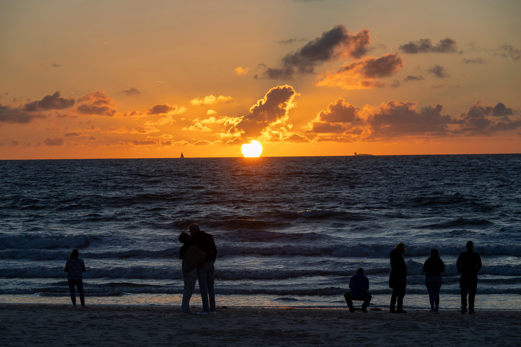 Sonnenuntergang an der Nordseeküste von Texel | Silhouetten von Spaziergängern und einem sich umarmenden Paar stehen am Sandstrand von Texel und blicken auf die Nordsee, während die Sonne am Horizont untergeht. Ein orange-goldener Himmel mit Wolkenformationen sowie leichte Wellen prägen die abendliche Küstenlandschaft der niederländischen Wattinsel. Aufnahme für Themen wie Tourismus, Reise, Erholung an der Nordsee, Abendstimmung und Naturerlebnis. - Realisiert mit Pictrs.com