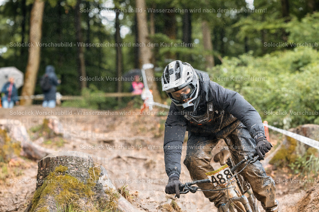 Mountainbike-Abenteuer | Ein Mountainbiker in voller Ausrüstung rast einen steilen, schlammigen Hang hinunter. Er trägt einen weißen Helm, eine dunkle Brille und eine schwarze Jacke und Hose. Sein Mountainbike ist mit einem Nummernschild versehen, das die Nummer 488 zeigt. Im Hintergrund sind Bäume und andere Mountainbiker zu sehen. - Realisiert mit Pictrs.com