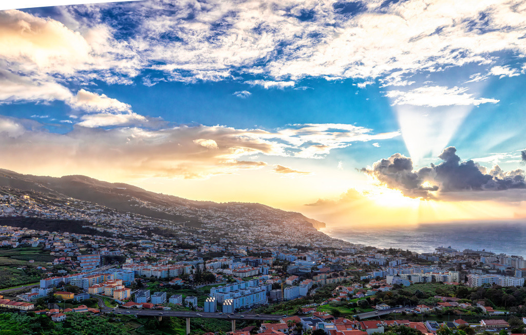 Spektakulärer Sonnenaufgang über Madeira, Funchal | Dieses Bild zeigt den Pico dos Barcelos in Funchal auf der Insel Madeira. Es handelt sich um einen Aussichtspunkt auf etwa 355 Metern über dem Meeresspiegel. Von hier aus hat man einen weiten Panoramablick über die Bucht von Funchal und das Stadtgebiet. Der Ort ist bekannt für seine spektakulären Sonnenuntergänge und Ausblicke auf den Atlantik. Die Umgebung wurde kürzlich neu gestaltet und bietet nun auch Grünflächen, Restaurants und Geschäfte mit lokalem Kunsthandwerk.  - Realisiert mit Pictrs.com