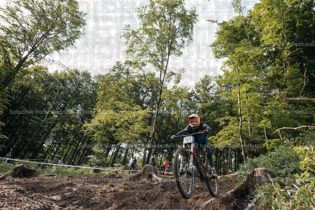 Mountainbike-Action im Wald | Ein Mountainbiker in voller Schutzausrüstung, inklusive Helm und Goggles, springt über eine Reihe von Baumstämmen in einem dichten Wald. Er trägt die Nummer 161 auf seinem Rücken. Im Hintergrund sind weitere Mountainbiker und eine Reihe von weißen Bändern zu sehen, die vermutlich die Strecke markieren. Die Szene vermittelt ein Gefühl von Geschwindigkeit und Abenteuer. - Realisiert mit Pictrs.com