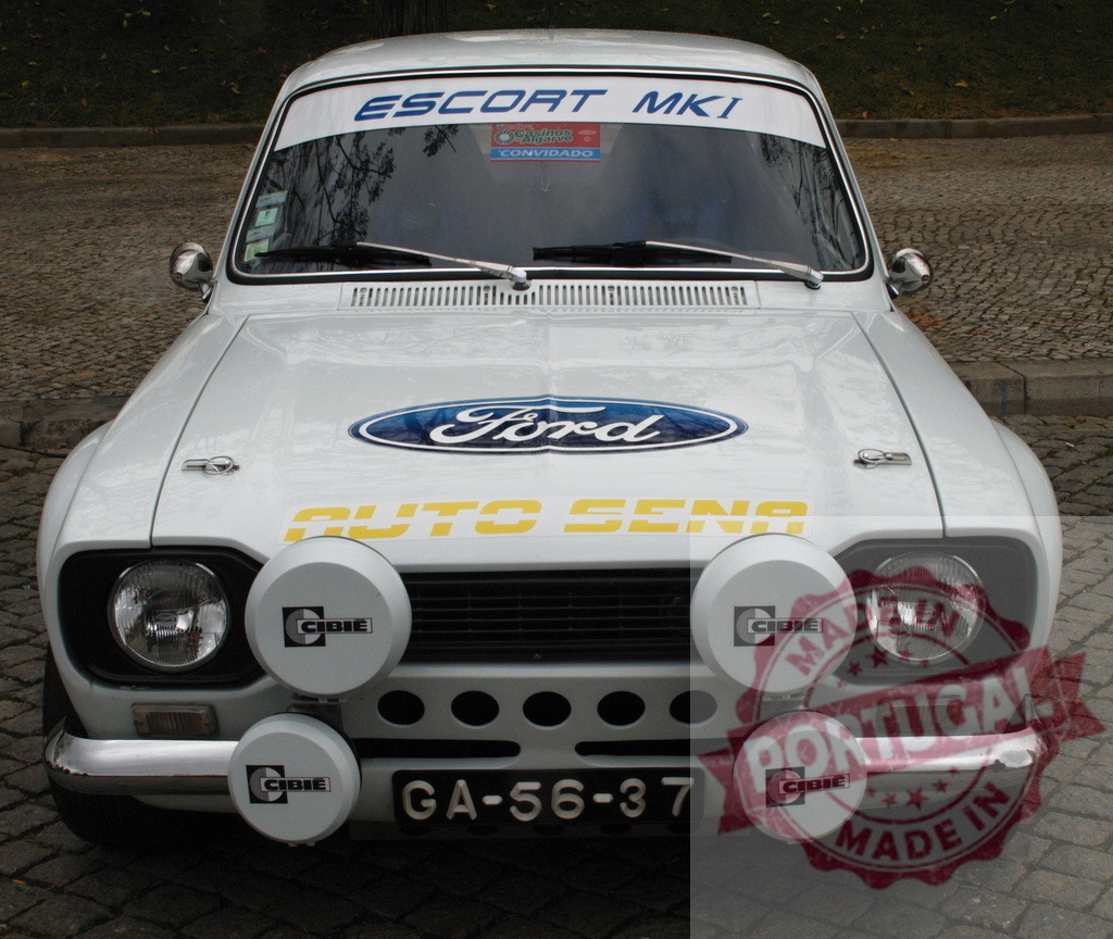 White Ford Escort MKI showing its front, parked on a cobblestone street | Front view of a white Ford Escort MKI rally car parked on a cobblestone street, featuring Cibie auxiliary lights and Auto Sena branding - Realized with Pictrs.com