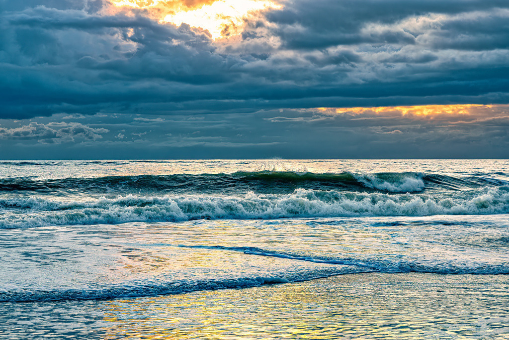 Sonnenuntergang am Strand von Westerland,Sylt,Wellen--Meer | Das Bild zeigt eine Meereslandschaft bei Sonnenuntergang am Strand von Westerland auf SyltDetails der SzeneHimmel: Dunkle, schwere Wolken verdecken den Großteil des Himmels, während ein goldener Lichtschimmer im oberen Zentrum durchbricht.Meer: Eine breite Welle bricht sich im mittleren Bildbereich und erzeugt weißen Gischt.Vordergrund: Der nasse Sandstrand reflektiert das warme, goldene Licht des Himmels. - Realisiert mit Pictrs.com
